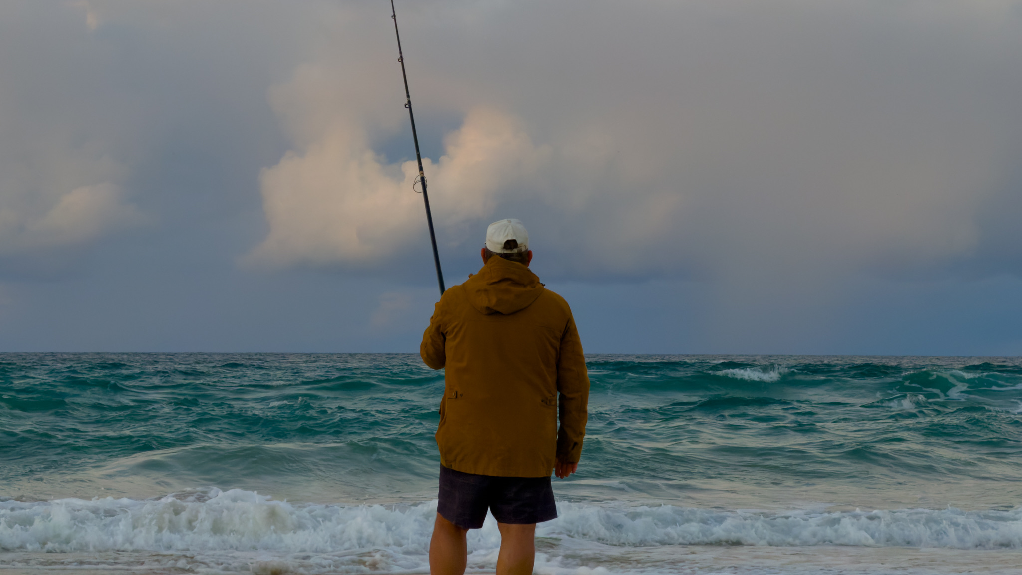 A beach fisherman casting into the swell at Currumbin under a moody sky.