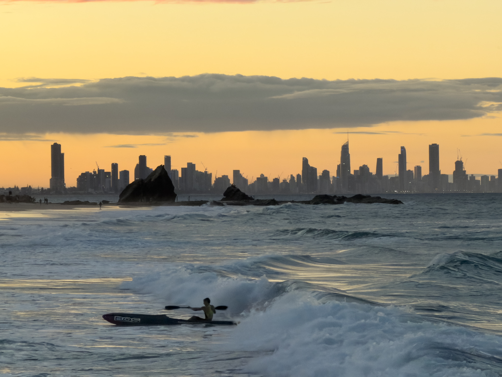 A surf-ski paddler on a wave at Currumbin, the rock and Gold Coast skyline glowing at sunset.