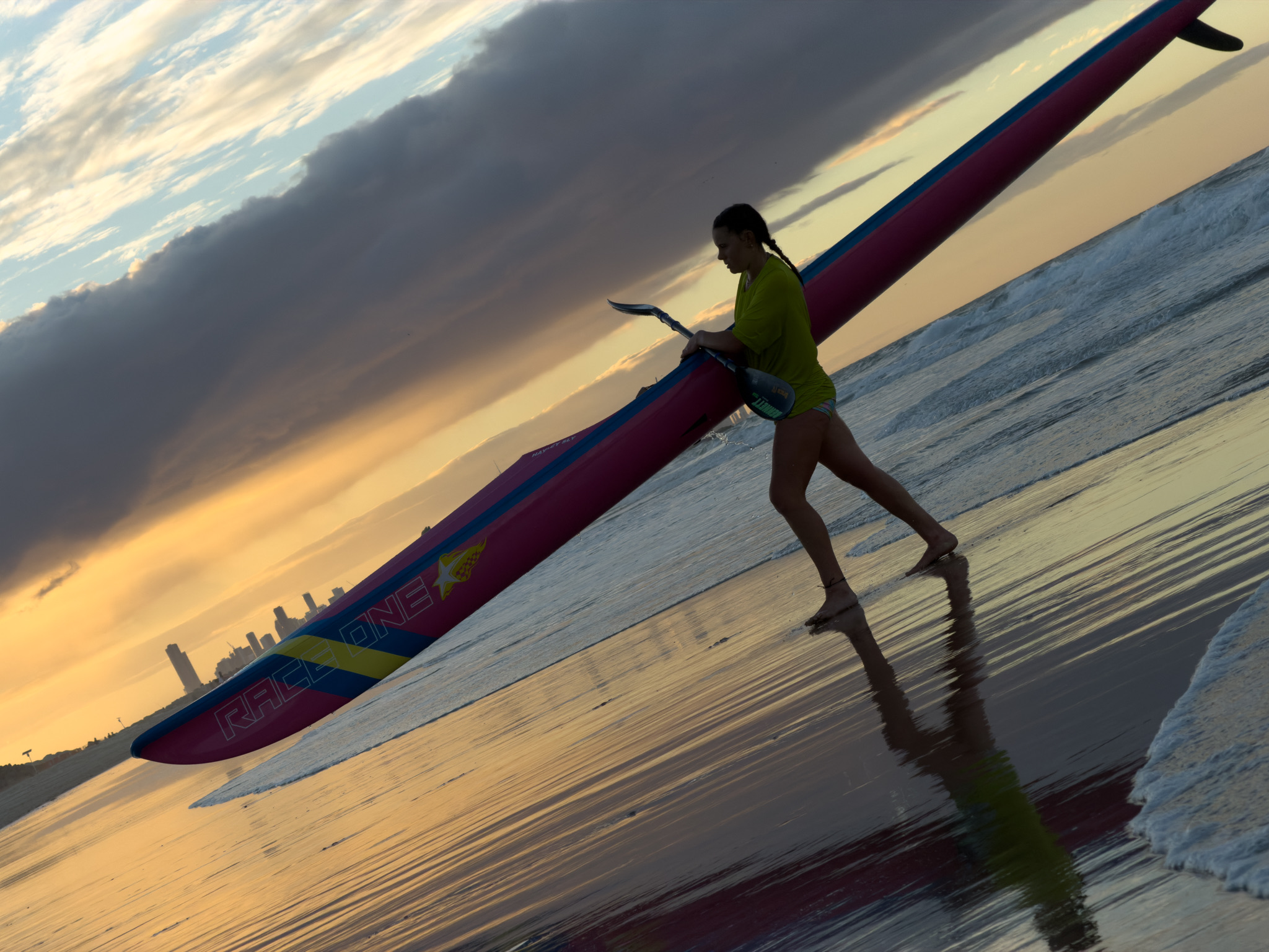 A surf-ski paddler carrying her ski to the water at Currumbin, the Gold Coast skyline behind at golden hour.