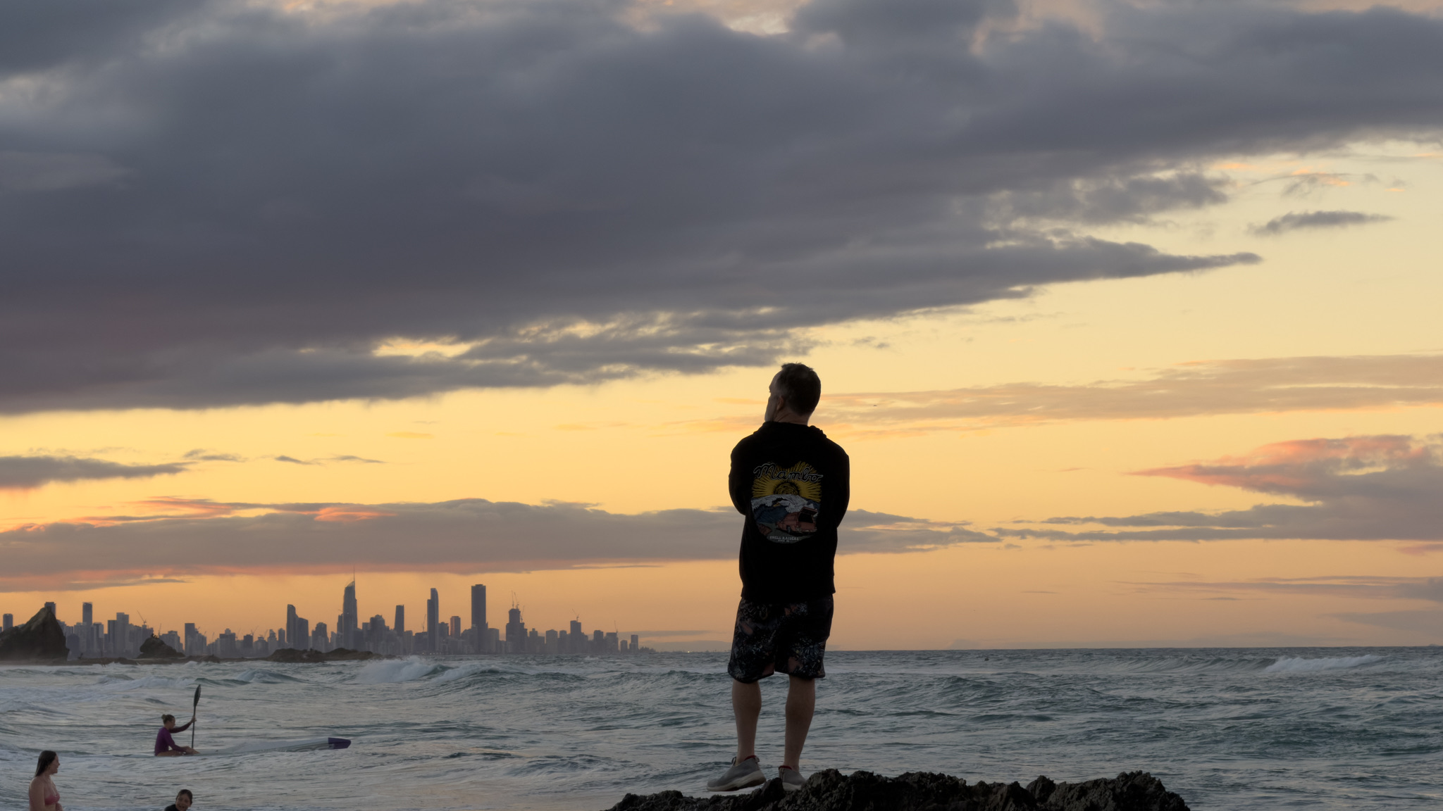 A figure on the rocks at Currumbin watching the surf, the Gold Coast skyline on the horizon.